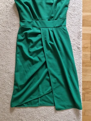 Grön festklänning  - Snygg grön kortklänning med omlottdetalj framtill och bred markerad midja. Klänningen har en enkel, elegant design med en ärm och är tillverkad i ett mjukt tyg som ger en smickrande passform. Använd en gång.