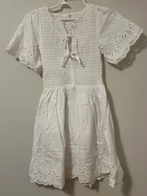 Vit kort klänning - Söt vit kortklänning med broderade spetsdetaljer på ärmar och nederkant. Klänningen har korta ärmar, smockad överdel och knytning fram. Perfekt för sommarens alla tillfällen. 