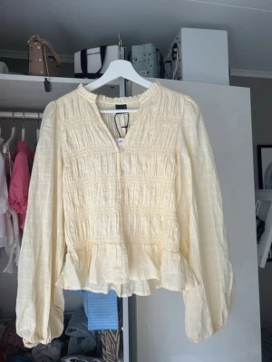 Gul smockad blus med ballongärm - Ljusgul blus med smockade detaljer och ballongärmar. Blusen har v-ringning och lätt volang nedtill, vilket ger en avslappnad men trendig look. Tillverkad i ett tunt, luftigt material som passar perfekt till vår och sommar.