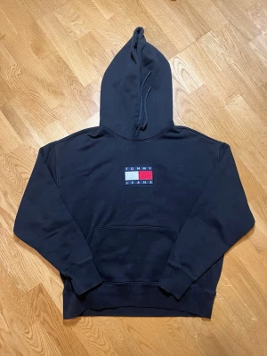 Svart hoodie Tommy Hilfiger - Säljer denna svarta hoodie från Tommy Jeans i storlek M med klassisk logga i vitt, rött och blått broderat på bröstet. Hoodien har huva med dragsko och en stor magficka. Perfekt för dig som gillar streetwear och vill ha en stilren look.  Vid frågor, extra bilder eller liknande är det bara att kontakta mig!