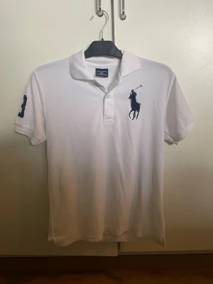 Vit pikétröja från Ralph Lauren - Klassisk vit pikétröja från Ralph Lauren med mörkblåt broderat logotyp på bröstet och nummerdetalj på ärmen. Tröjan har krage, knappar framtill och korta ärmar. Perfekt till en avslappnad men ändå stilren look.