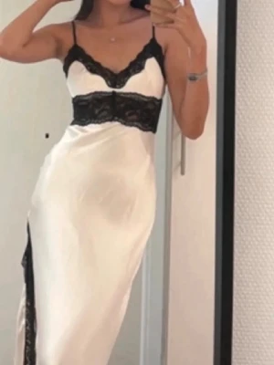 Vit långklänning med svart spets - Säljer en elegant vit långklänning med tunna axelband och svarta spetsdetaljer vid byst och midja. Klänningen har slits på sidan och öppen rygg, vilket ger en snygg och modern look. Tillverkad i ett glansigt material som känns lyxigt.