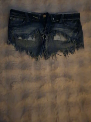 Mörkblå jeansshorts med fransar - Säljer ett par mörkblå jeansshorts med råa, fransiga kanter och slitningar framtill. Shortsen har klassisk femficksdesign, bälteshällor och knappgylf. Perfekta för en avslappnad och trendig sommarlook.