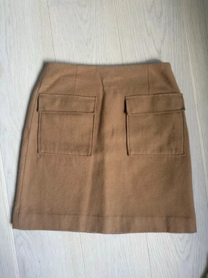 Kort höstkjol i ull - Snygg beige/brun kortkjol med två stora fickor framtill. Kjolen har en enkel och stilren design som passar till många olika outfits. Passar en size small
