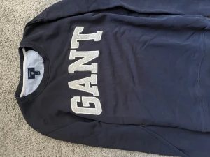 Mörkblå sweatshirt från GANT - Mörkblå sweatshirt från GANT med stora vita bokstäver framtill. Klassisk rund halsringning och mjukt material, perfekt för en avslappnad stil. Snygg och enkel att matcha med jeans eller joggers.