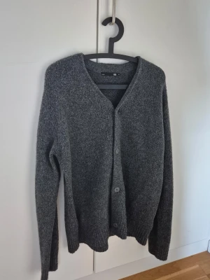 Stickaf Cardigan - Mysig grå stickad cardigan med v-ringning och knäppning framtill. Långa ärmar och ribbade muddar vid ärmslut och nederkant. Perfekt för lager på lager och enkel att matcha med olika outfits.
