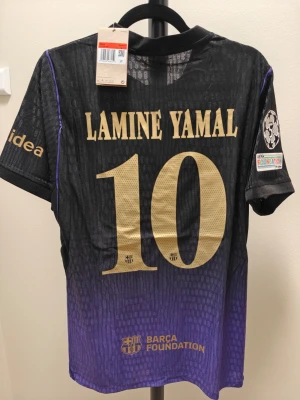 Barça spelar version Lamine Yamal matchtröja svart/lila - Snygg FC Barcelona matchtröja i svart och lila med guldiga detaljer och nummer 10 Lamine Yamal på ryggen. Tröjan har officiella klubbmärken, Champions League-märke på ärmen och sponsorloggor. Tillverkad i lätt och ventilerande funktionsmaterial.