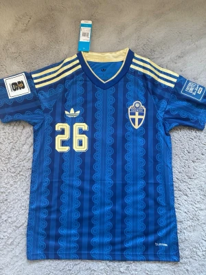 Sverige bortatröja 2026 - Säljer en blå fotbollströja från Adidas med Sveriges landslagsemblem, nummer 26 och namnet Ali på ryggen. Tröjan har gula detaljer, V-ringning och klassiska Adidas-ränder på axlarna. Tillverkad i ett lätt och ventilerande material, perfekt för match eller träning.