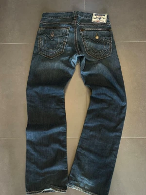 True Religion blå jeans  - Säljer ett par blå jeans från True Religion med tydliga kontrastsömmar och stora fickor bak med lock och dekorativa knappar. Klassisk rak passform och patch med logga bak i midjan. Perfekta för dig som gillar streetwear och detaljerade jeans.