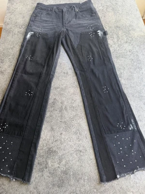Svarta jeans med nitar och råa kanter - Svarta jeans i denim med raka ben, dekorerade med små silvriga nitar och slitna detaljer. Jeansen har råa, fransiga benslut och klassisk femficksmodell. Perfekt för dig som gillar en trendig och edgy vibe.