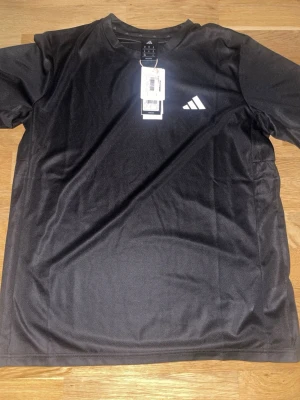 Svart tränings-tshirt från Adidas - Svart sportig t-shirt från Adidas med klassisk vit logga på bröstet. Tillverkad i ett lätt och ventilerande funktionsmaterial som passar perfekt för träning och aktivitet. Rund halsringning och korta ärmar för en bekväm passform.
