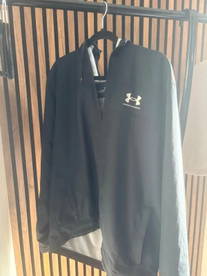 Svart vindjacka från Under Armour - Snygg svart vindjacka från Under Armour med dragkedja och vit logga på bröstet. Jackan har huva, meshfoder och praktiska fickor. Perfekt för blåsiga dagar och sportiga outfits.