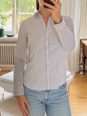 Vit och blårandig skjorta - Klassisk vit skjorta med tunna blå ränder och lång ärm. Skjortan har knappar framtill och en smickrande, något figurnära passform. 