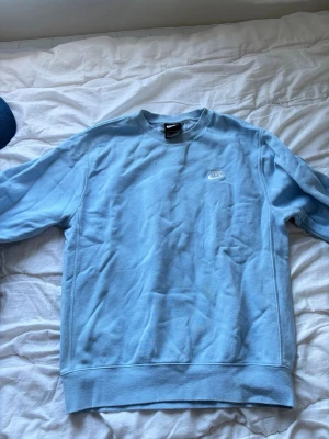 Ljusblå sweatshirt från Nike - Säljer en ljusblå sweatshirt från Nike med klassisk rund halsringning och broderad vit Nike-logga på bröstet. Tröjan har ribbade muddar vid ärmslut och nederkant. Perfekt för en avslappnad och sportig stil. En liten missfärgning, som knappt syns