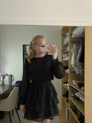 Svart prickig volangklänning - Söt svart klänning med vita prickar och volangkjol. Klänningen har långa, transparenta ärmar och rund halsringning. Kjolen är i två lager med volanger för extra rörelse. Perfekt för dig som gillar en lekfull och trendig stil.