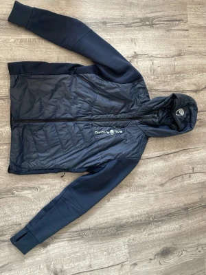Mörkblå vindjacka Sail Racing - Snygg mörkblå vindjacka från Sail Racing med huva och hel dragkedja framtill. Jackan har släta ärmar med tumhål och quiltad kropp i lättviktigt material. Perfekt för blåsiga dagar och sportiga outfits.