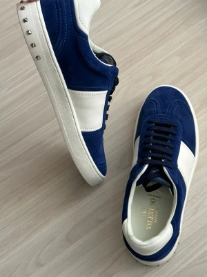 Blå och vita sneakers från Valentino - Snygga sneakers från Valentino i blå mocka och vitt läder. Skorna har svarta skosnören, vita sulor och coola nitar på hälen. Perfekta för dig som vill ha en stilren men ändå unik look.