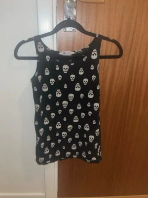 Svart linnetopp med dödskallemönster H&M - Cool svart linnetopp från H&M med vita dödskallar över hela tyget. Perfekt för dig som gillar edgy stil! Toppen är ärmlös och har en rund halsringning. Materialet känns mjukt och stretchigt, troligen bomull eller bomullsblandning.