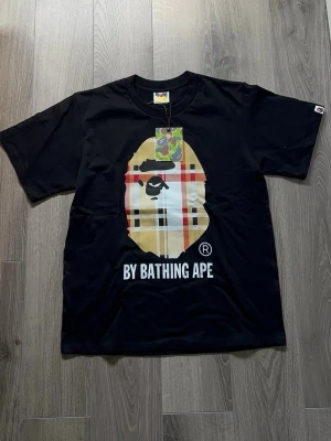 Svart BAPE t-shirt  - Svart t-shirt från BAPE med den klassiska apansiluetten i rutig design på bröstet och texten 'BY BATHING APE' under. T-shirten har rund hals och korta ärmar. Perfekt för dig som gillar streetwear och vill sticka ut.
