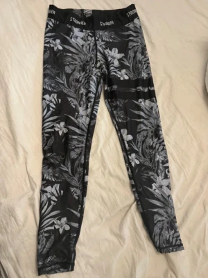 Svarta blommiga leggings från Stronger - Snygga svarta leggings från Stronger gamla kollektion med grått och vitt blommönster. Hög midja med elastisk linning där det står 'STRONGER'. Tillverkade i en stretchig mix av polyester och elastan, perfekta för träning eller chill.
