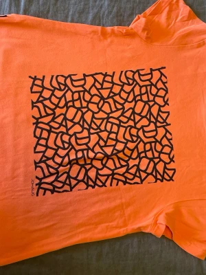 Orange t-shirt från Johaug NOW stl.XS - Stl.XS, Snygg orange t-shirt från Johaug NOW i bomull med ett svart abstrakt tryck på framsidan. Släppt sömn nere pp ryggen runt 1 cm (syns på bild 3-4)