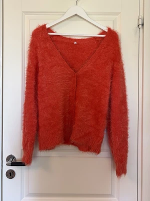 Fluffig korallröd kofta med v-ringning - Supermjuk och fluffig kofta i en härlig korallröd färg. Koftan har v-ringning, knäppning framtill med matchande knappar och långa ärmar. Materialet är lurvigt och ger en mysig känsla. Perfekt för dig som vill ha något extra cozy i garderoben.