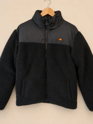 Svart teddyjacka Ellesse y2k retro  - Mysig svart teddyjacka från Ellesse med hög krage och dragkedja framtill. Överdel i slätt material och nederdel i fluffigt teddytyg. Liten broderad logga på bröstet och två sidofickor. Perfekt för kalla dagar och riktigt snygg streetstil.