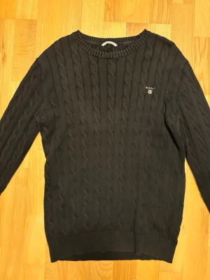 Svart kabelstickad tröja från Gant - Kabelstickad svart tröja från Gant i premium bomull. Klassisk rund halsringning, ribbade muddar och diskret Gant-logga på bröstet. Perfekt för dig som gillar stilrena och tidlösa plagg med snygg struktur.