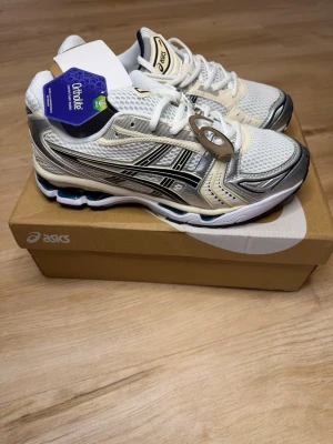 Asics Gel-Kayano 14 vita/silver  - Snygga Asics Gel-Kayano 14 sneakers i vitt och silver med svarta detaljer. Ovandel i mesh och syntet för bra andningsförmåga och komfort. Klassisk retrodesign med Ortholite-innersula och stötdämpande sula. Perfekta för dig som gillar sportig stil.