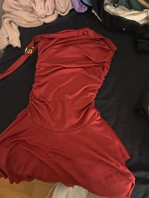 Röd one shoulder klänning - Snygg röd one shoulder klänning i ett glansigt material med draperade detaljer och en dekorativ axelrem med gulddetalj. Kort modell som ger en trendig och elegant look. Perfekt för dig som vill sticka ut.