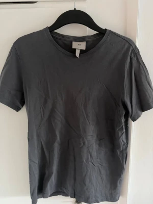 Mörkgrå basic t-shirt från H&M - En enkel mörkgrå t-shirt från H&M med rund halsringning och korta ärmar. Tillverkad i mjuk bomull som känns skön mot huden. Perfekt för en avslappnad och stilren look.