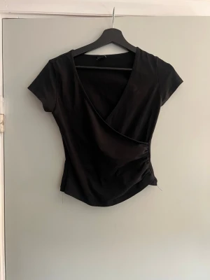 Svart omlottopp med rynkade detaljer - Snygg svart omlottopp med korta ärmar och v-ringning. Toppen har rynkade detaljer i sidan som ger en schysst form och sitter tajt. Perfekt att styla med jeans eller kjol för en clean look.