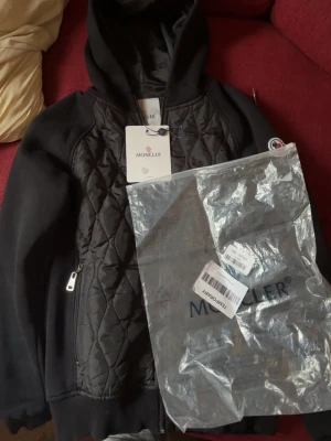 Svart quiltad jacka från Moncler - Svart jacka från Moncler med quiltad front, huva och dragkedja. Jackan har Moncler-logga på ärmen och ribbade muddar. Perfekt för dig som vill ha en stilren och exklusiv look.