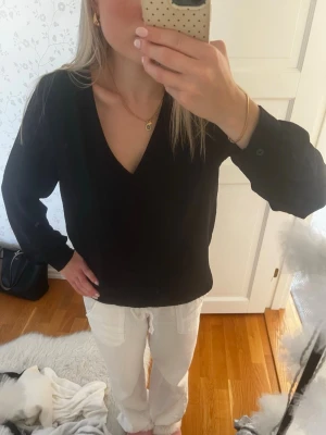 Svart blus med v-ringning - Stilren svart blus med djup v-ringning och lång ärm. Blusen har en lätt textur i tyget och en avslappnad passform som ger en snygg siluett. Perfekt att matcha med både jeans och kjol för en trendig look.