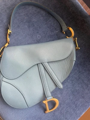 Blå Dior Saddle Bag i läder  - Säljer en ikonisk blå Dior Saddle Bag i läder med guldfärgade metalldetaljer och det klassiska D-spännet. Väskan har en unik sadelform och justerbart axelband. Insidan är fodrad i blå mocka och har en innerficka med dragkedja.