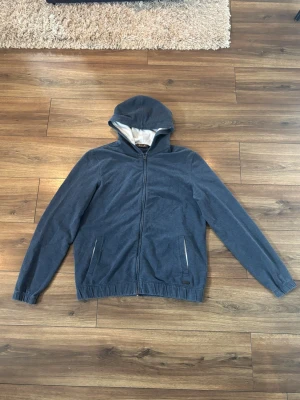 Loro Piana Zip Hoodie - Snygg mörkgrå hoodie ifrån Loro Piana med dragkedja framtill och två fickor. Insidan av huvan är ljusare och materialet känns mjukt och bekvämt, perfekt för chill dagar. Resår vid ärmslut och nederkant ger en skön passform. Liten detalj med metallbricka vid fickan. 