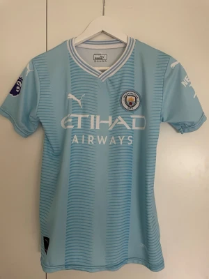 Manchester City matchtröja Puma - Ljusblå Manchester City matchtröja från Puma med klubbmärke, Etihad Airways-tryck och Premier League-logga. Tröjan har vita detaljer, korta ärmar och Haaland 9 på ryggen. Perfekt för fotbollsfans och träning.