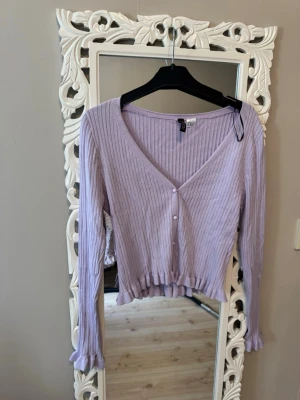 Ljuslila ribbad kofta från H&M - Supersöt ljuslila kofta från H&M i ribbad trikå med v-ringning och knäppning framtill. Modellen är croppad med långa ärmar och volangkant nedtill och vid ärmslut. Perfekt för att addera färg till din outfit!