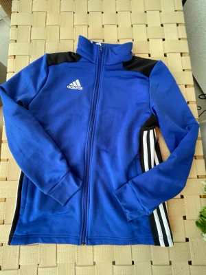 Blå Adidas träningsjacka  - Snygg blå träningsjacka från Adidas med dragkedja framtill, vita ränder på sidorna och svarta detaljer vid axlar och sidor. Jackan har hög krage och lång ärm, perfekt för träning eller uppvärmning. Adidas-logga på bröstet.