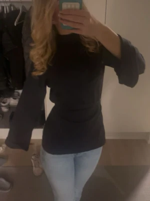 Svart topp med vida ärmar - Snygg svart topp med hög hals och vida långa ärmar. Modellen är figurnära i midjan och har en enkel, stilren design utan mönster eller detaljer. Perfekt att matcha med jeans eller kjol för en trendig look.