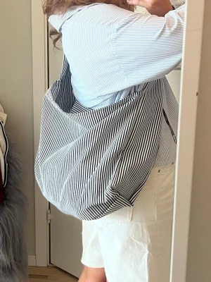 Randig svart axelväska i tyg - Säljer en stor axelväska i svart tyg med tunna vita ränder. Väskan har en bred axelrem och rymlig form, perfekt för att bära med sig allt man behöver. Enkel och stilren design som passar till många outfits.