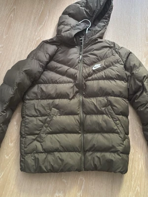 Olivgrön pufferjacka från Nike - Säljer en olivgrön pufferjacka från Nike med huva och dragkedja framtill. Jackan har två sidofickor med knappar och ett broderat Nike-märke på bröstet. Perfekt för kalla vinterdagar och riktigt skön att bära.