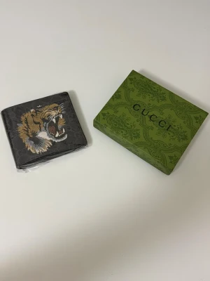 Svart plånbok med tigertryck - Snygg svart plånbok från med ett stort broderat tigerhuvud på framsidan. Plånboken har diskret GG-mönster i bakgrunden och är tillverkad i tåligt material. Kommer med original grön box.
