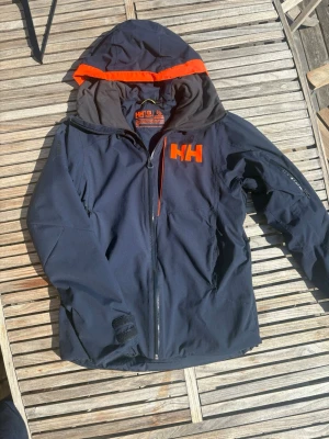 Mörkblå  Helly Hansen vindjacka med huva - Snygg mörkblå vindjacka från Helly Hansen med orange detaljer och stor logga på bröstet. Jackan har huva, dragkedja framtill och justerbara muddar. Perfekt nu till hösten men även för skidåkning. Materialet är slitstarkt och vattenavvisande.