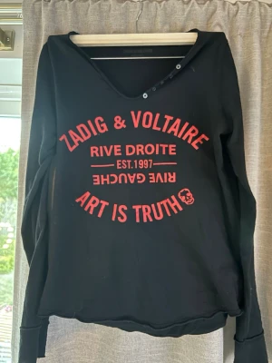 Svart tröja Zadig & Voltaire - Cool svart långärmad tröja från Zadig & Voltaire med röd textprint framtill: 'Art is Truth', 'Rive Droite' och 'Est. 1997'. Tröjan har en djupare rund halsringning med knappdetaljer och avslappnad passform.