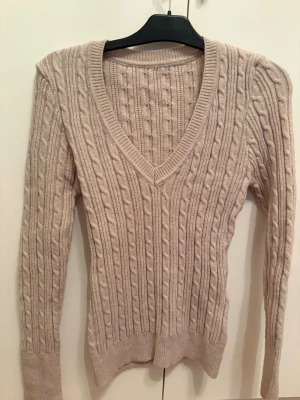 Beige kabelstickad tröja V-ringad - Väldigt stretchig material, den är storlek L men KAN passa S-M. Säljer en beige kabelstickad tröja med djup V-ringning och långa ärmar. Tröjan har ribbade muddar vid ärmslut och nederkant, samt ett klassiskt kabelmönster över hela plagget. Perfekt för dig som gillar en enkel men snygg stil.
