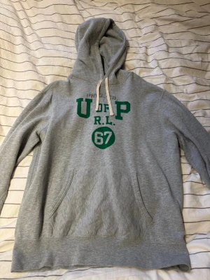 Grå hoodie från Polo Ralph Lauren - Snygg grå hoodie från Polo Ralph Lauren med grön text och siffran 67 på bröstet. Klassisk känguruficka framtill och justerbar huva med vita snören. Tillverkad i mjukt bomullsmaterial, perfekt för en avslappnad streetwear-look.