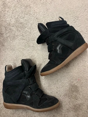 Svarta Isabel Marant skor - Säljer ett par svarta wedge sneakers från Isabel Marant med cool design och kardborreband. Skorna har en hög siluett och är gjorda i mocka med detaljer i läder samt en brun gummisula. Perfekta för dig som vill ha en edgy och trendig look.