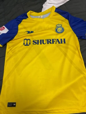 Gul och blå Al Nassr fotbollströja - Säljer en gul och blå Al Nassr fotbollströja med 'SHURFAH' tryck på bröstet och Ronaldo 7 på ryggen. Tröjan har klubbmärke på bröstet, korta ärmar och är tillverkad i ett lätt, ventilerande material perfekt för en varm dag  