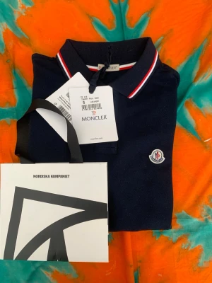 Mörkblå piké från Moncler, S - Säljer en mörkblå pikétröja från Moncler i storlek S. Pikén har klassisk krage med vita och röda ränder, samt Monclers ikoniska logga på bröstet. Tillverkad i bomull för en skön och luftig känsla. Perfekt till jeans eller shorts!
